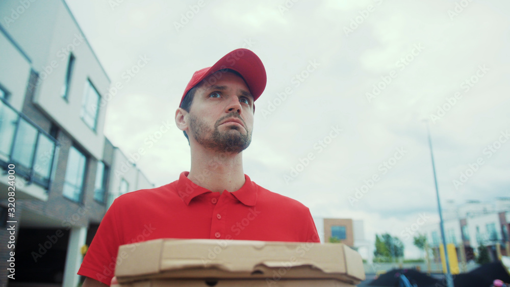 Low angle portrait young courier holding pizza boxes delivering food order. Close up tired delivery man is lost looking for right house in big modern courtyard. Home delivery services. Low angle portrait young courier holding pizza boxes delivering food order. Close up tired delivery man is lost looking for right house in big modern courtyard. Home delivery services.
