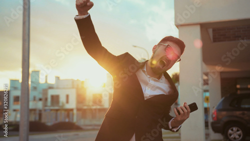 Attractive satisfied young man clenching fist jumping with happiness at sunset. Outdoor portrait happy businessman i proud of victory smiling and having fun in the street. photo