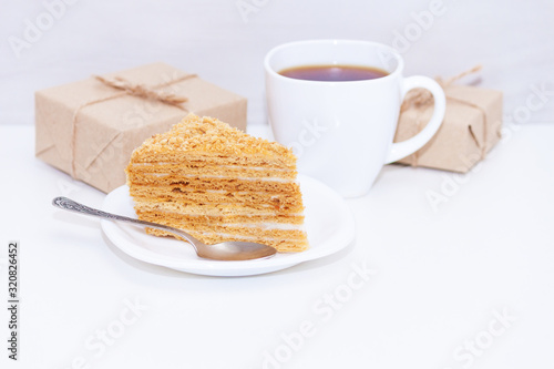 Slice of homemade layers honey cake, cup of coffe, present box or gift on wooden white background. Happy morning, copy space.