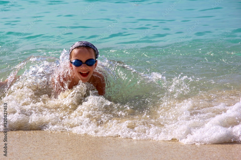 Laughing little cute boy in transparent splashes of rushing wave. Happy eight year old boy enjoys splashing waves in shallow sea water on exotic tropical beach.  We play, relax. Summer active leisure.