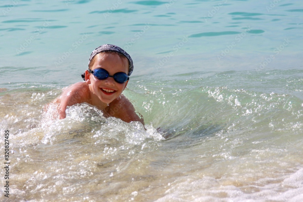 Smiling little cute boy in transparent splashes of rushing wave. Happy eight year old boy enjoys splashing waves in shallow sea water on exotic tropical beach.  We play, relax. Summer active leisure.