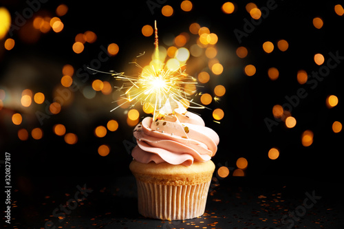 Delicious birthday cupcake with sparkler on black table against blurred lights