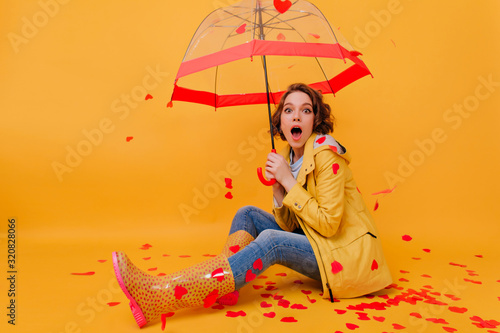 Surprised enthusiastic girl in rubber autumn shoes sitting on the floor. Studio portrait of glad female model posing, surrounded by paper hearts.