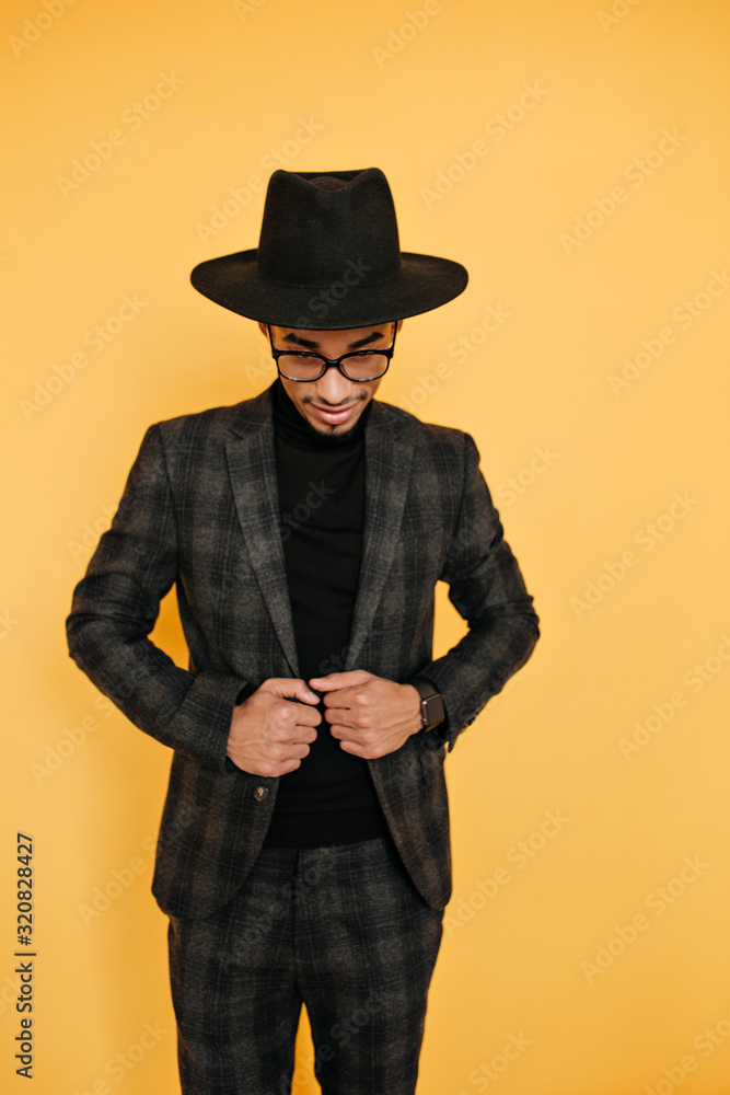 Slim african man in big black hat posing in elegant suit. Indoor photo of glad black guy in gray attire isolated on yellow background. Slim african man in big black hat posing in elegant suit. Indoor photo of glad black guy in gray attire isolated on yellow background.