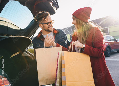A cute young couple at the parking in front of a shopping mall, packing bags into a car trunk.