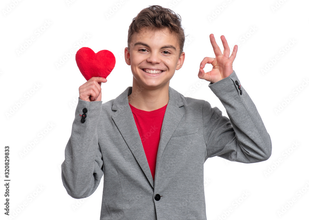 Happy Valentines Day. Cute young Teen Boy in Love with Red plush Heart in his hands. Smiling Child in grey suit making Ok Gesture and looking at camera, isolated on white background. Happy Valentines Day. Cute young Teen Boy in Love with Red plush Heart in his hands. Smiling Child in grey suit making Ok Gesture and looking at camera, isolated on white background.