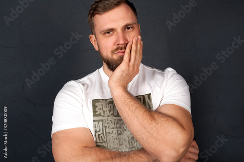 A man with a thoughtful face  with a serious look  holding his face with his hand  in a white t-shirt on a gray background. The concept of the issues or emotions and life problems.