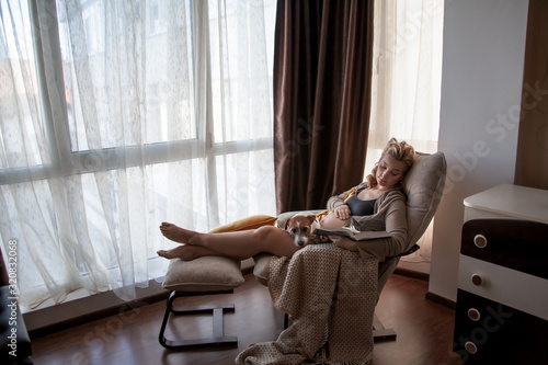 Pregnant woman siting on cozy armchair with cup of tea at home infront of the window. The owner of pet expecting baby and relaxing with dog . Hygge concept.