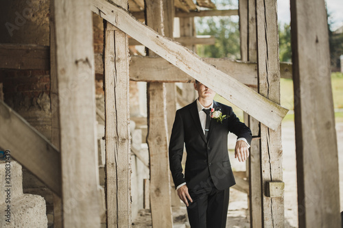 Fashion portrait of a groom outside. Half of his face is hidden behind a wooden plank