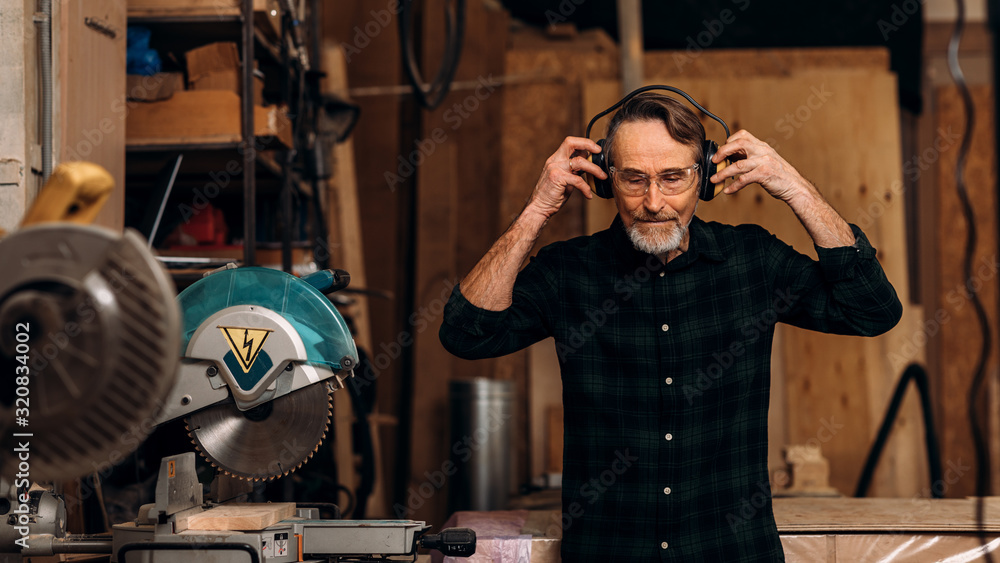 Senior carpenter wearing noise-canceling headphones preparing for working on saw machine