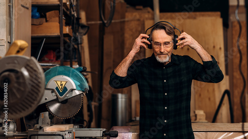 Senior carpenter wearing noise-canceling headphones preparing for working on saw machine