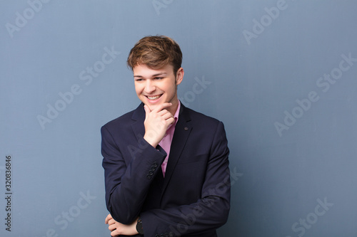 young blonde man smiling with a happy, confident expression with hand on chin, wondering and looking to the side isolated against flat wall photo