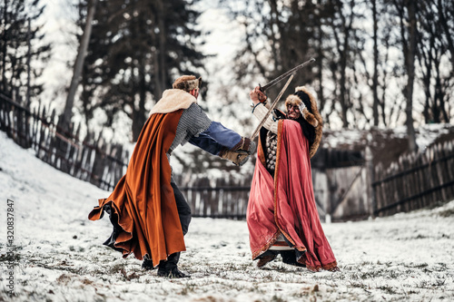 Battle of two knights in colored robes and hats near forest and wooden fortress. Knights fight in winter, in the snow