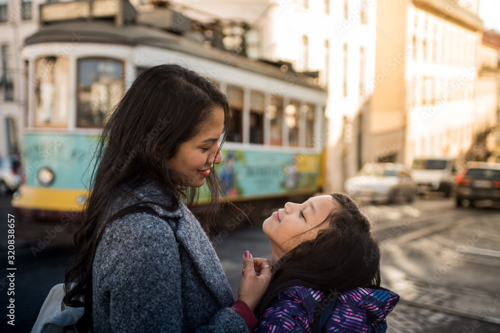 Latin mother and daughter looking affectionately outside on a Lisbon street, in the background there is a classic tram from Portugal