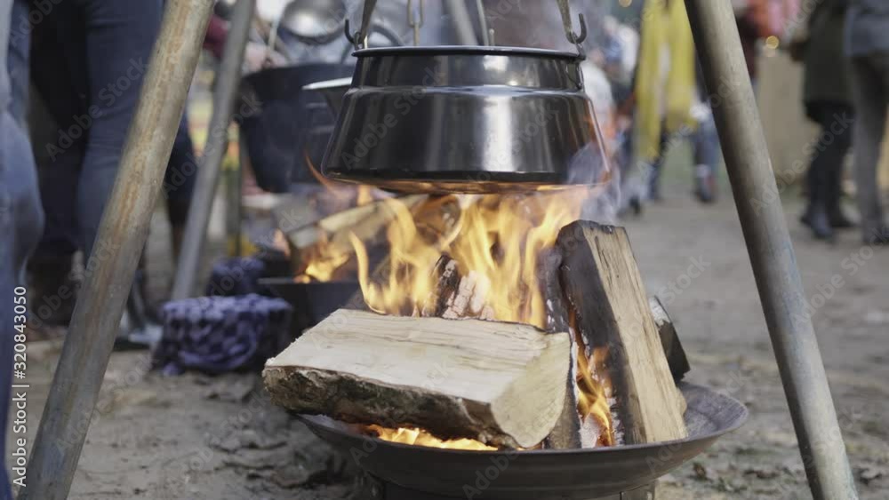A stove hanging on a wooden tripod above a campfire in place with many people standing around it. A stove hanging on a wooden tripod above a campfire in place with many people standing around it.