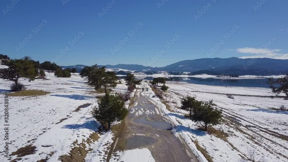 People are walking on a snowy footpath surrounded by pine trees near lake in winter People are walking on a snowy footpath surrounded by pine trees near lake in winter