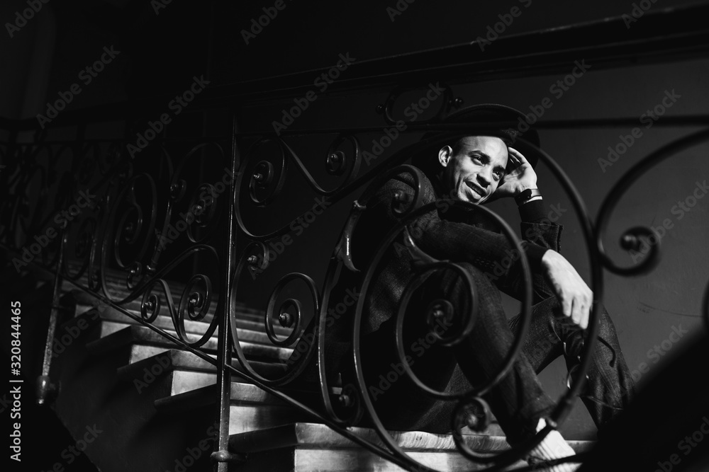 Obraz premium Male model posing in the dark. Indoor black and white portrait of inspired african man sitting on the steps.