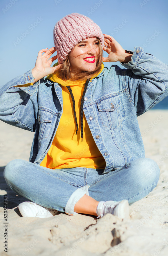 Young beautiful woman in denim clothes and knit hat sitting on sand by the sea