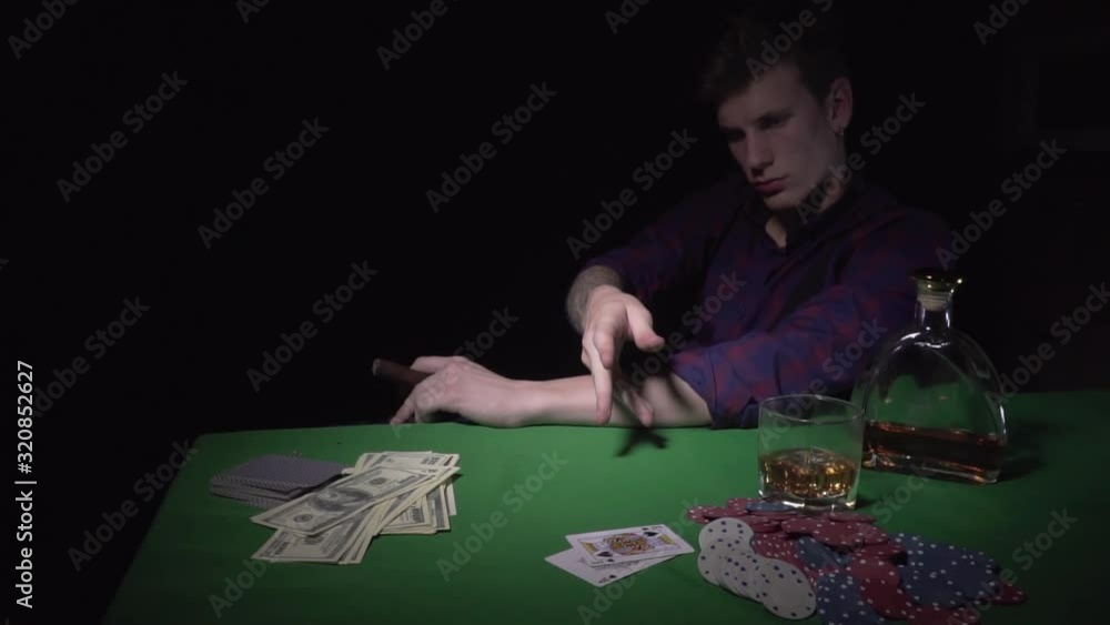 A young man with a cigar at a card table in a casino. A guy throws cards on a green table in a casino. On the table is a bottle of alcohol, a glass and playing chips. Moving camera. Slow motions A young man with a cigar at a card table in a casino. A guy throws cards on a green table in a casino. On the table is a bottle of alcohol, a glass and playing chips. Moving camera. Slow motions