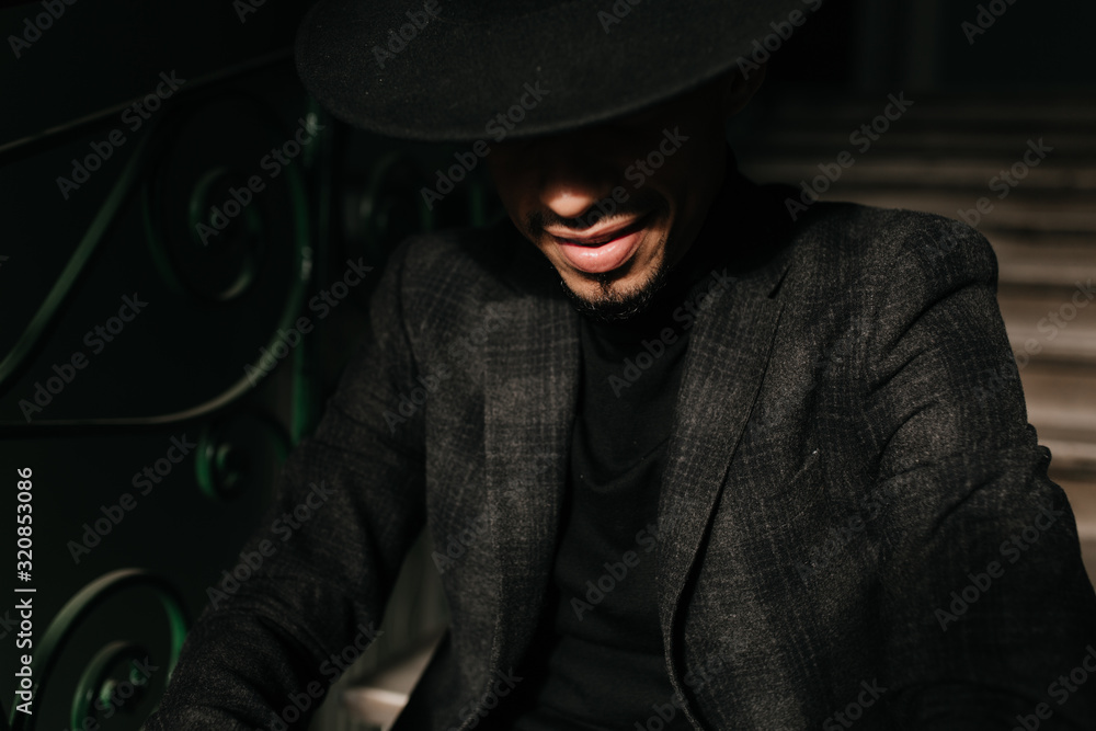 Obraz premium Close-up portrait of african man in formal wear posing in the dark. Refined male model in big hat sitting on stairs.