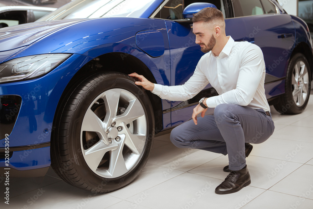 Full length shot of a handsome elegant man examining wheels and tires of a car he is buying at the dealership Full length shot of a handsome elegant man examining wheels and tires of a car he is buying at the dealership