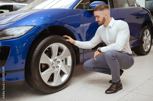 Full length shot of a handsome elegant man examining wheels and tires of a car he is buying at the dealership © Ihor