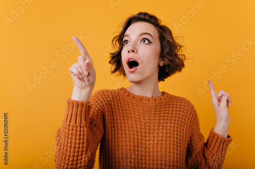 Close-up portrait of enthusiastic girl with suprised face expression looking around. Studio shot of lovable curly lady isolated on orange background.