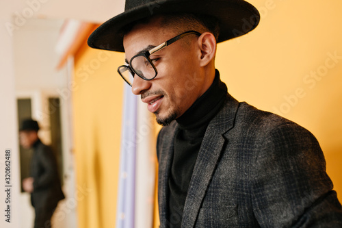 Bearded african man in hat posing in studio. Indoor close-up portrait of thoughtful black guy in glasses standing near yellow wall.