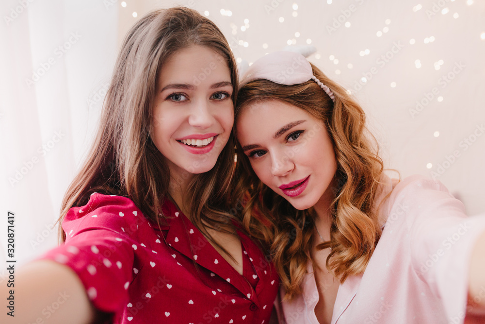 Dark-haired girl expressing positive emotions and making selfie in morning. Pretty curly woman in pink eyemask spending time with sister. Dark-haired girl expressing positive emotions and making selfie in morning. Pretty curly woman in pink eyemask spending time with sister.