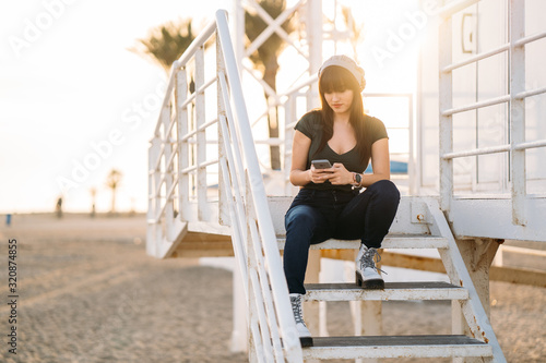 Beautiful young woman sitting on a the lifeguard booth using smart phone at beach during sunset