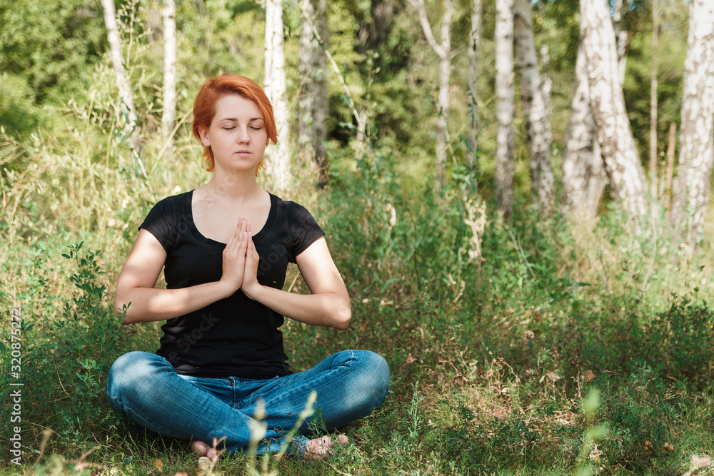 Fototapeta premium Freedom and joyful concept with copy space. Girl teenager meditates in park. Red haired woman sitting on grass.
