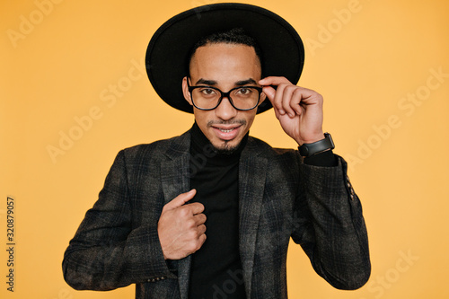 Studio portrait of handsome brown-eyed male model playfully touching his hat. Indoor photo of well-dressed african man in good mood.