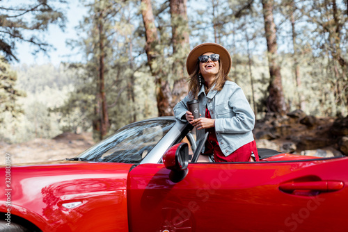 Young stylish woman enjoying beautiful views of nature while traveling on convertible car, standing on the roadside in the forest