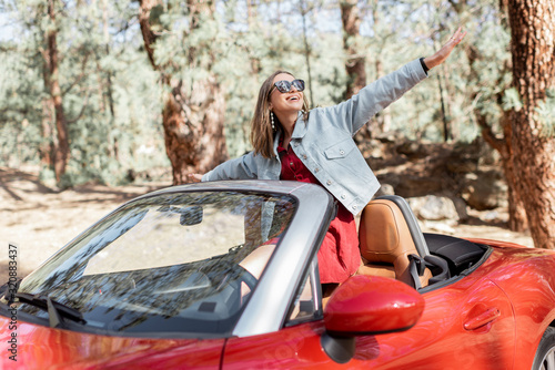 Young stylish woman enjoying traveling by convertible car on nature, pulling hands out of the car. Carefree lifestyle and travel concept