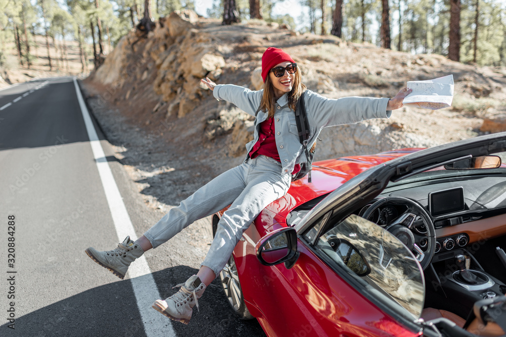 Lifestyle portrait of a carefree woman dressed casually in jeans and red hat sitting on the car hood, enjoying road trip on the mountain road