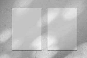 Two empty white vertical rectangle poster or business card mockups with diagonal dappled light spots on gray concrete wall. Flat lay, top view. For advertising, brand design, stationery presentation.