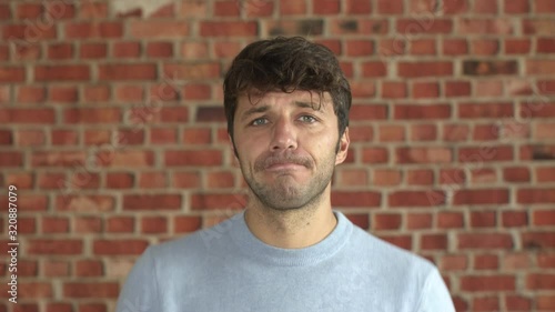 Suggestive portrait of handsome young man with light blue sweater shrugging undecided about a choice. Attractive careless bearded guy.