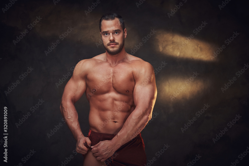 Pensive muscular bodybuilder is demonstrating his leg muscles at dark photo studio.