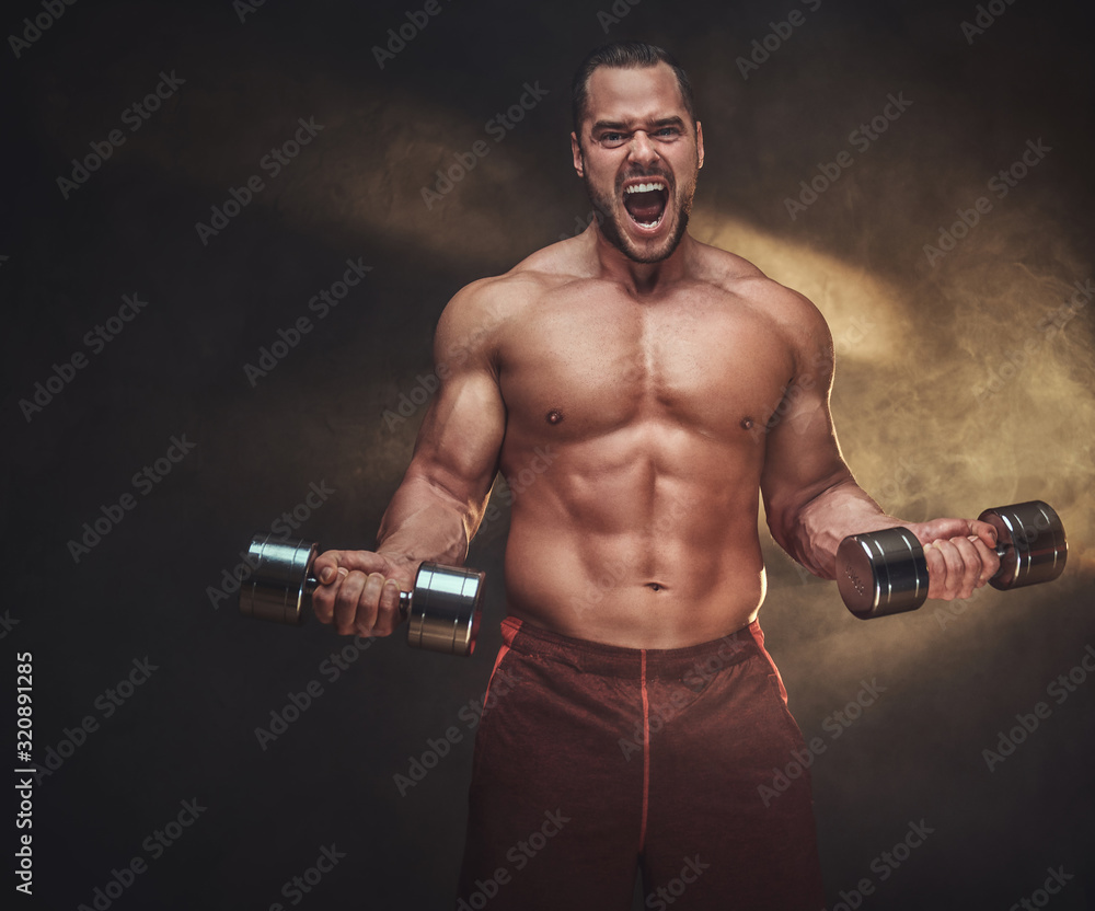 Shirtless muscular man is screaming while doing exercises with dumbbells.