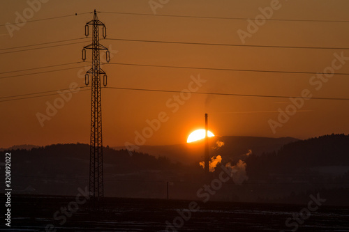 A sunset behind the heating plant. The plant releases steam. In front, the transmission tower stands. Looks like symbol of old, not environmentally friendly industry. 