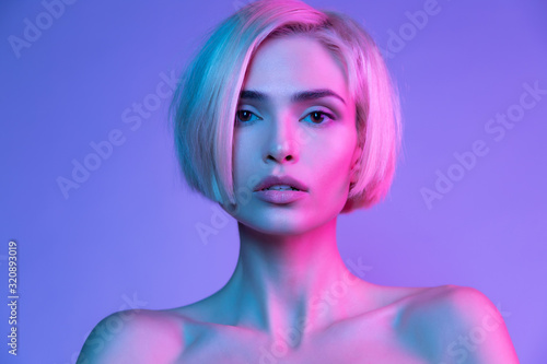 Gorgeous young woman with messy hair and naked shoulders looking at camera in neon portrait. Fashion studio headshot with awesome girl having natural makeup. Prefect beautiful female face