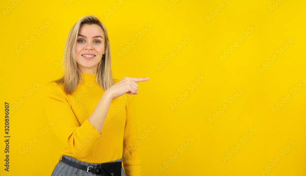 Amazing young woman points finger the side. Caucasian model smiling and looking in camera. Isolated on yellow background. Copy space Amazing young woman points finger the side. Caucasian model smiling and looking in camera. Isolated on yellow background. Copy space