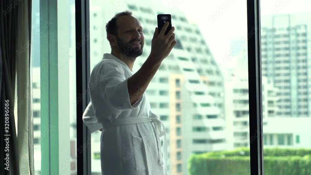 Young man having video chat on smartphone by window at home, slow motion Young man having video chat on smartphone by window at home, slow motion