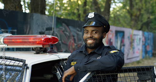 Portrait of handsome African American young policeman in uniform and cap. Front view of police officer smiling at camera outdoors. Cop on the background with police car and old lane.