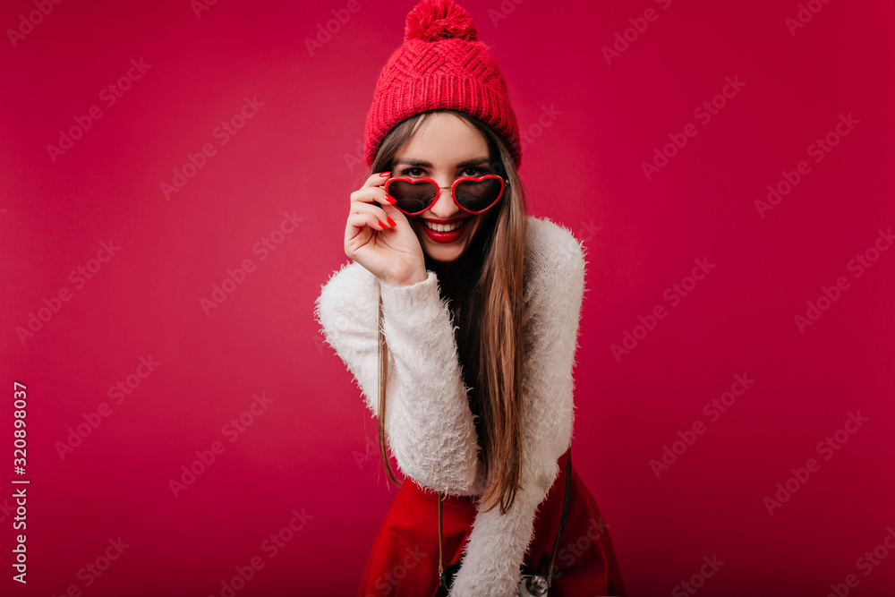 Smiling girl in cute winter hat playfully posing in studio. Indoor portrait of carefree long-haired lady in glasses standing on claret background and looking to camera. Smiling girl in cute winter hat playfully posing in studio. Indoor portrait of carefree long-haired lady in glasses standing on claret background and looking to camera.