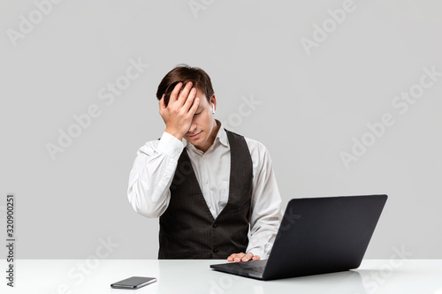 Man in a white t-shirt and grey vest is shocked looking at the laptop on a white table