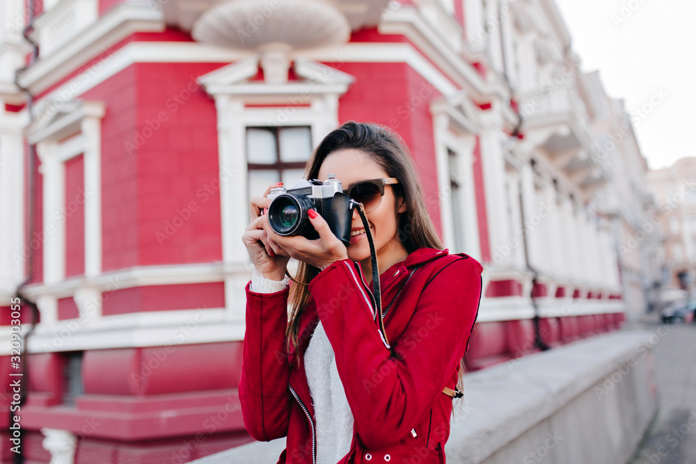 Fascinating female photographer working in morning in city. Outdoor photo of pleased young woman in red jacket and white shirt standing with camera near beautiful building.