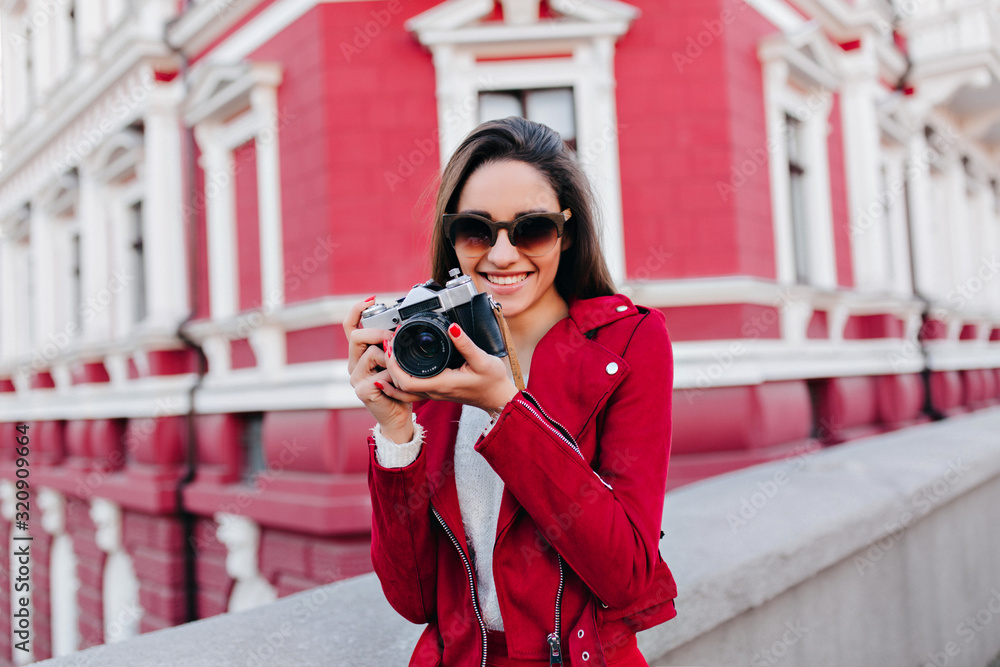 Inspired caucasian girl posing with happy smile after street photoshoot. Cheerful brunette woman with camera fooling around in weekend.
