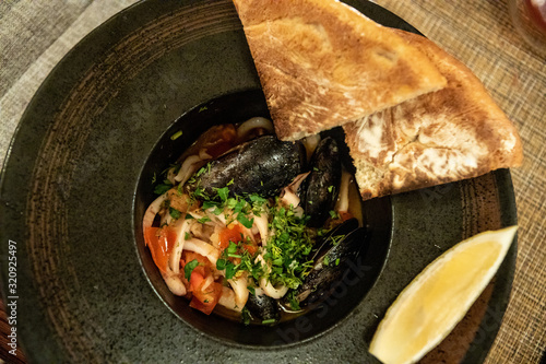 Roast mussels and squid with tomatoes and herbs in a dark plate with lemon and slices of bread. Delicacy