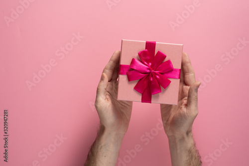 Fototapeta Naklejka Na Ścianę i Meble -  Valentine's Day celebration concept. A nice gift from a loved one. Box with a bow in male hands on a delicate pink background. Copy space. Flat lay.